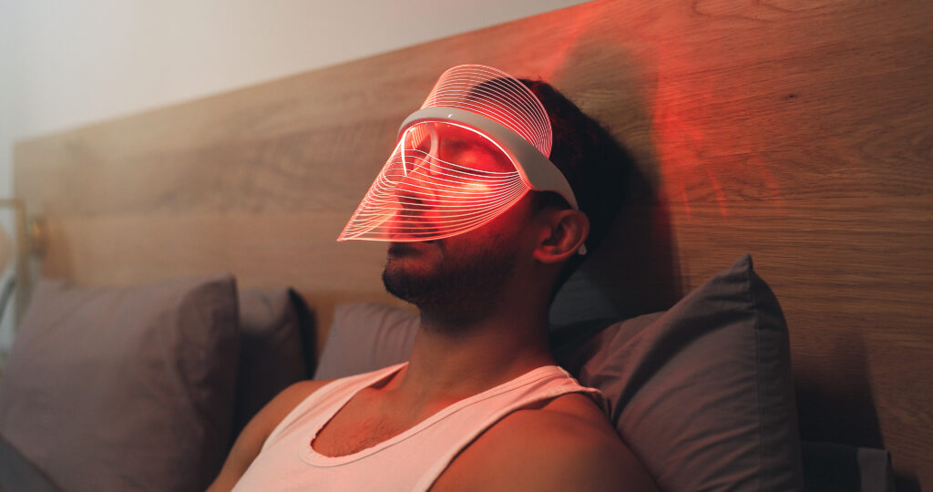 Man lying in bed wearing a red light therapy mask, using red light therapy for sleep before bedtime