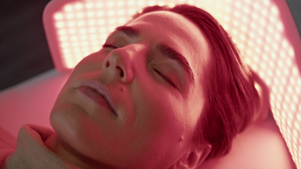 Woman relaxing under a red LED light panel, using red light therapy for sleep and skin recovery.