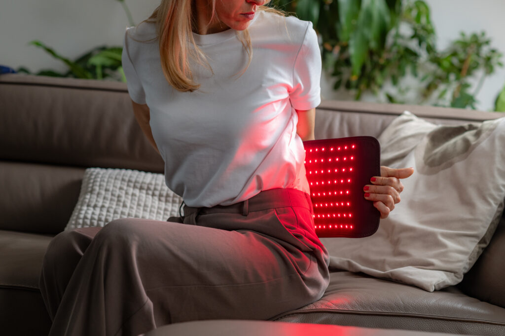 Woman using a portable red light therapy panel on her lower back while sitting on the couch.