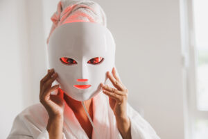 Woman using an at-home red light therapy LED mask over a towel and robe.